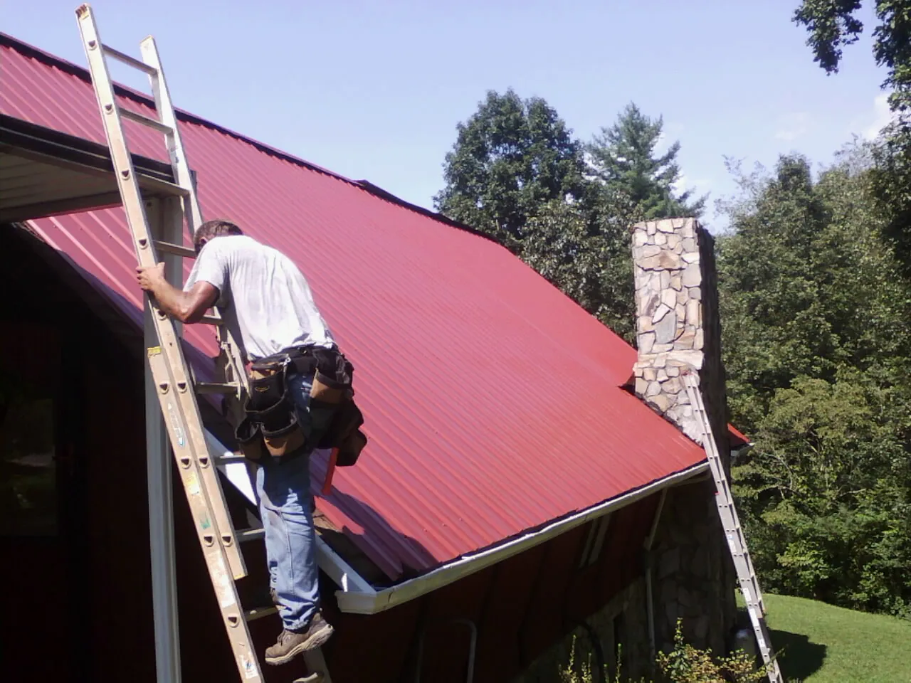 Roofer on a ladder beside a red metal roof and stone chimney — Soffit & Fascia Repair by Roofing Brothers in Helmetta